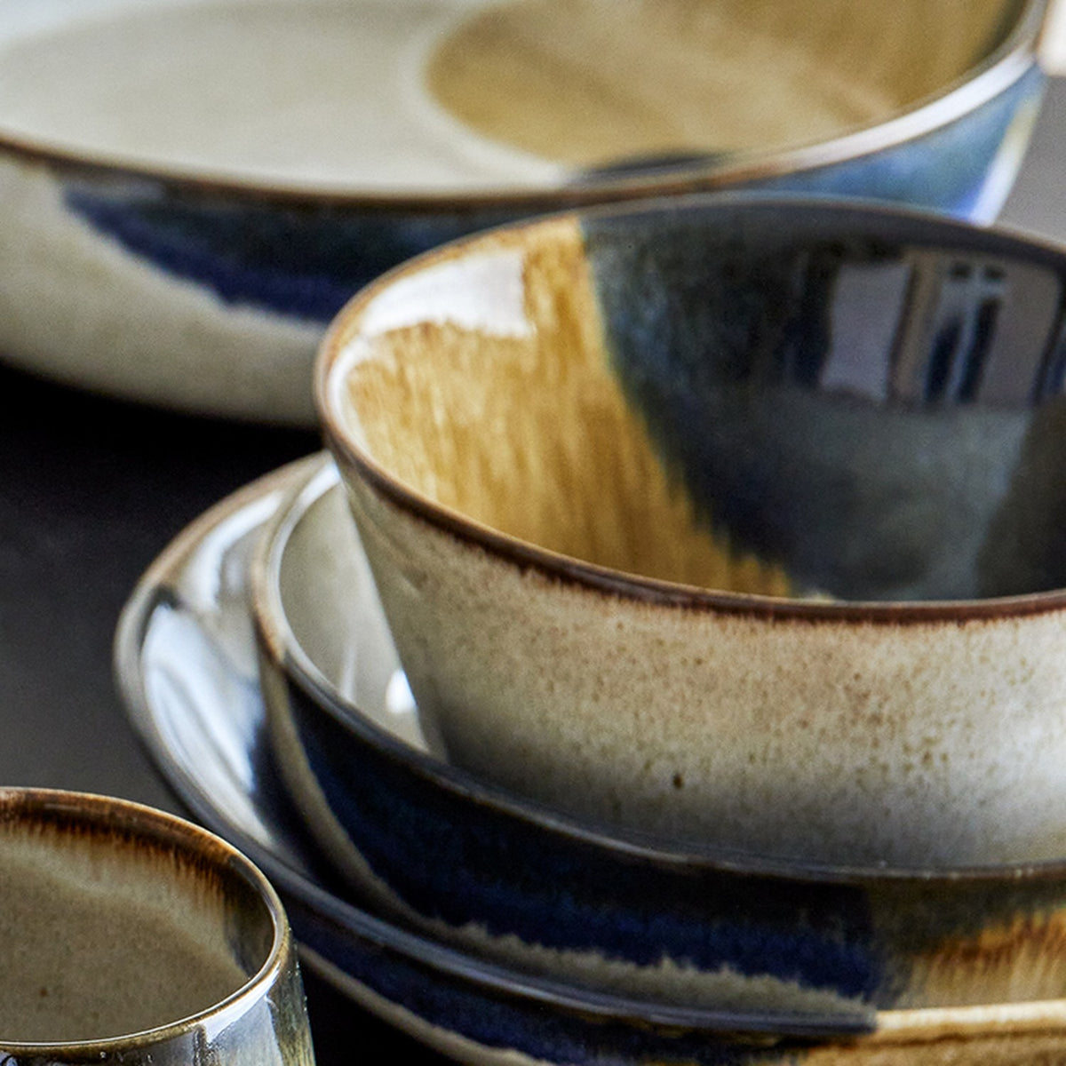 Jules Stoneware Bowl 13cm in Multi, shown among matching plates and a cup with a glossy reactive glaze blending blue, green and tan tones with drips over the rim.