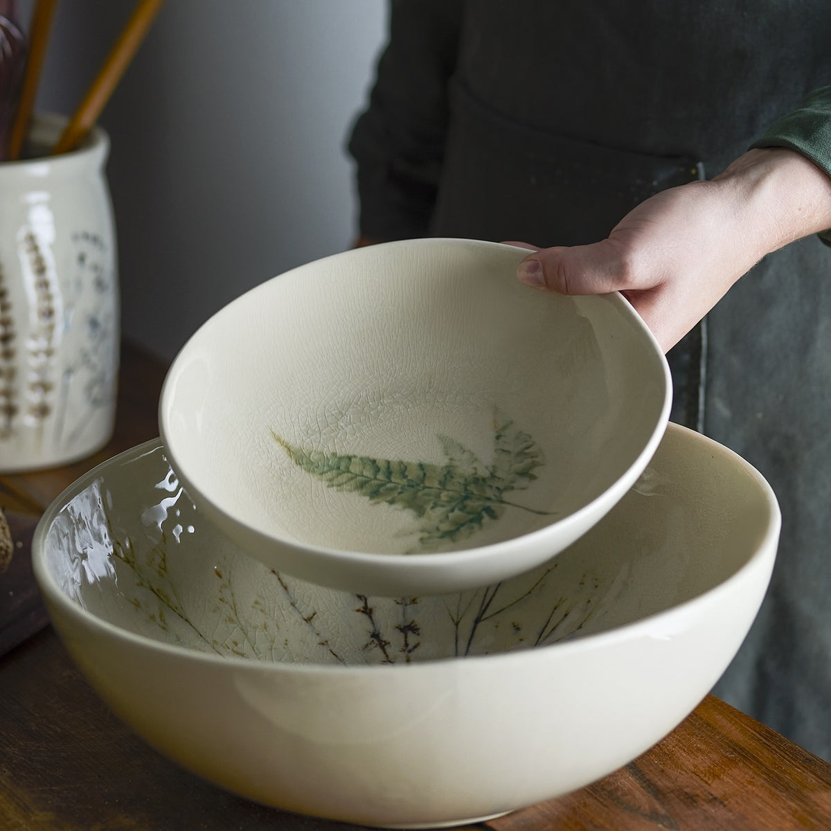 A hand holds the Bea Crackle Stoneware Deep Plate, cream with a fern leaf motif, above a larger matching bowl with botanical sprigs, on a wooden table.