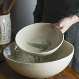 A hand holds the Bea Crackle Stoneware Deep Plate, cream with a fern leaf motif, above a larger matching bowl with botanical sprigs, on a wooden table.
