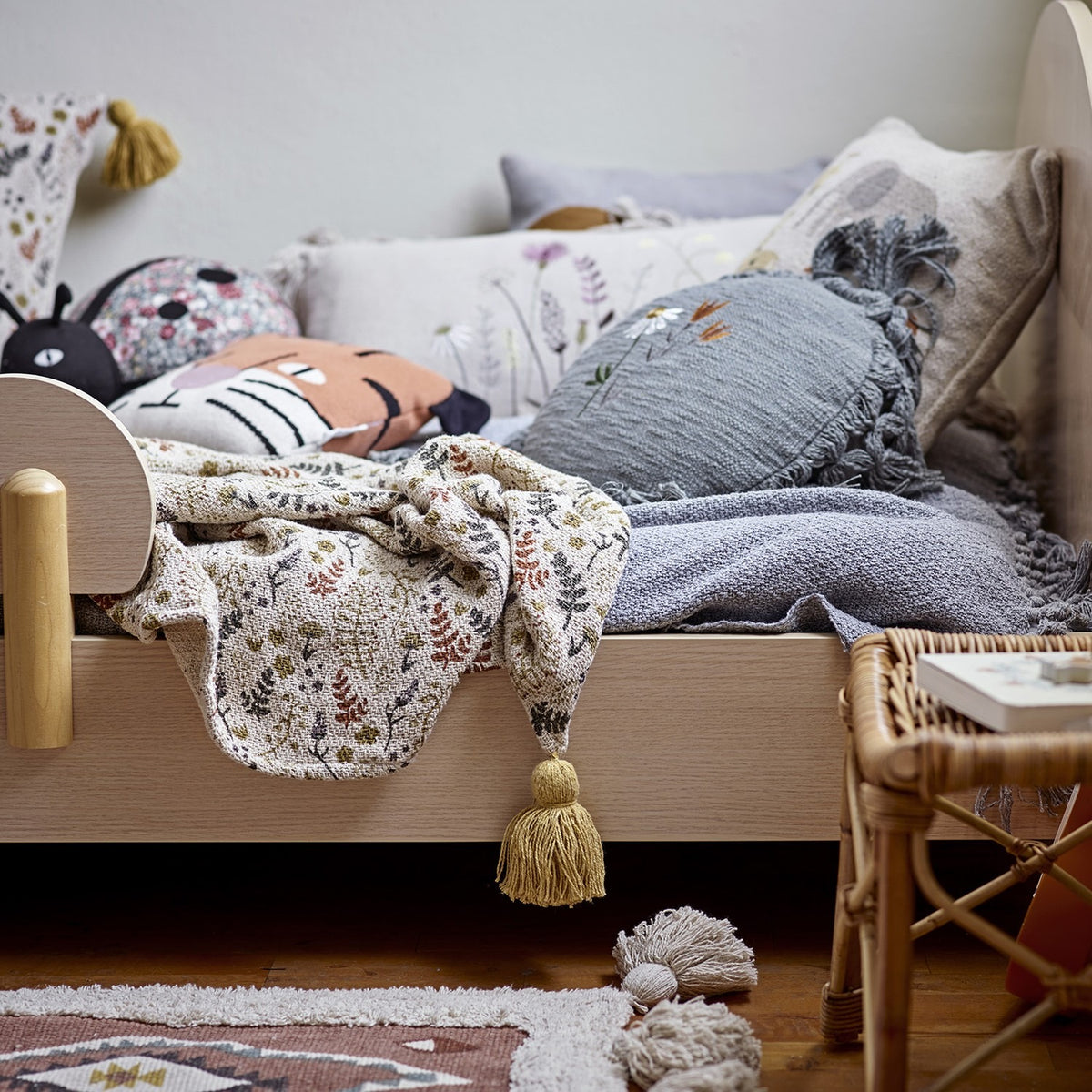 Filipa Floral Cotton Throw in multicoloured fern and floral print with corner tassels, draped over a wooden children's bed among cushions.