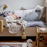 Filipa Floral Cotton Throw in multicoloured fern and floral print with corner tassels, draped over a wooden children's bed among cushions.