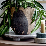 Nala Terracotta Plant Pot in Black with hand-painted leaf-like strokes, holding a green leafy plant on a stone tray beside a lit candle on marble holders.