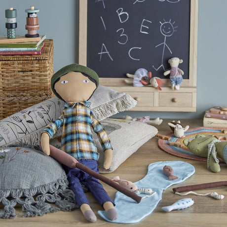 Sigge cotton and linen rag doll in a blue plaid shirt and navy trousers with knitted hat, seated among cushions in a playroom with a chalkboard and toys.