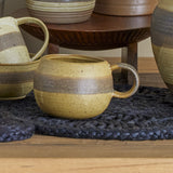 Solange Stoneware Mug 350ml in Green, rounded stoneware mug with speckled glaze and brown banding, loop handle, shown among matching ceramic pieces on a woven black mat.