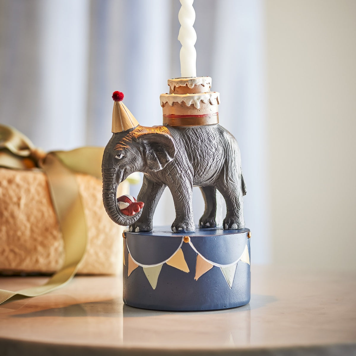 Multicoloured 18cm Flor resin candle holder shaped as an elephant in a party hat carrying a tiered cake, on a bunting-decorated base, shown indoors with a gift box nearby.
