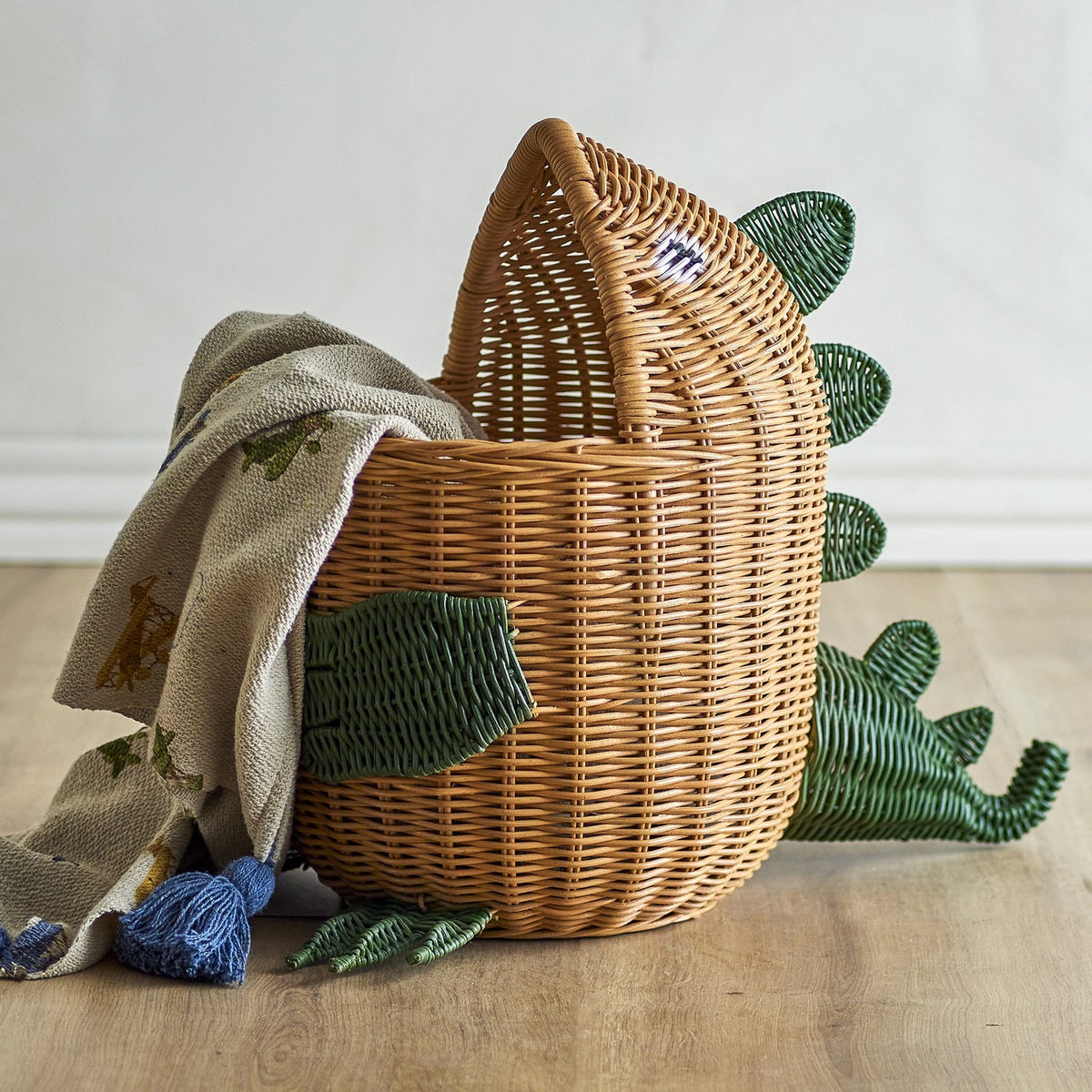 Eddi Dinosaur Rattan Basket in natural rattan with green woven spines, arm and clawed feet details, shown with a cloth draped over its head shape against a light background.