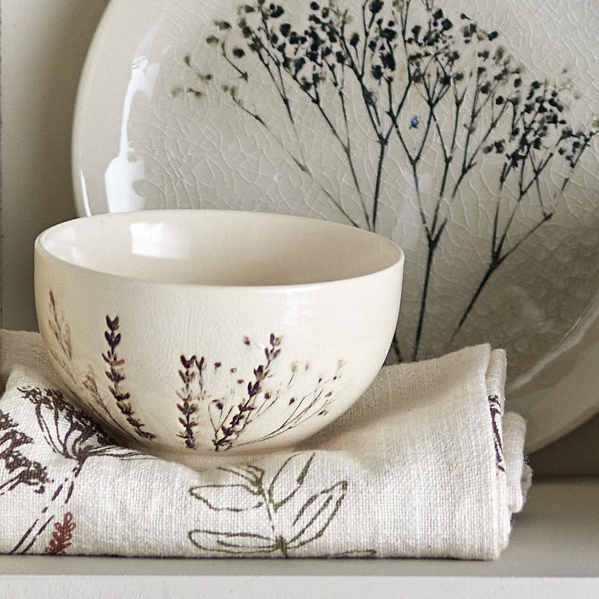 Bea Floral Stoneware Bowl 11.5cm in beige with sprig floral motif, plain cream interior, shown beside a patterned plate and floral linen napkin.