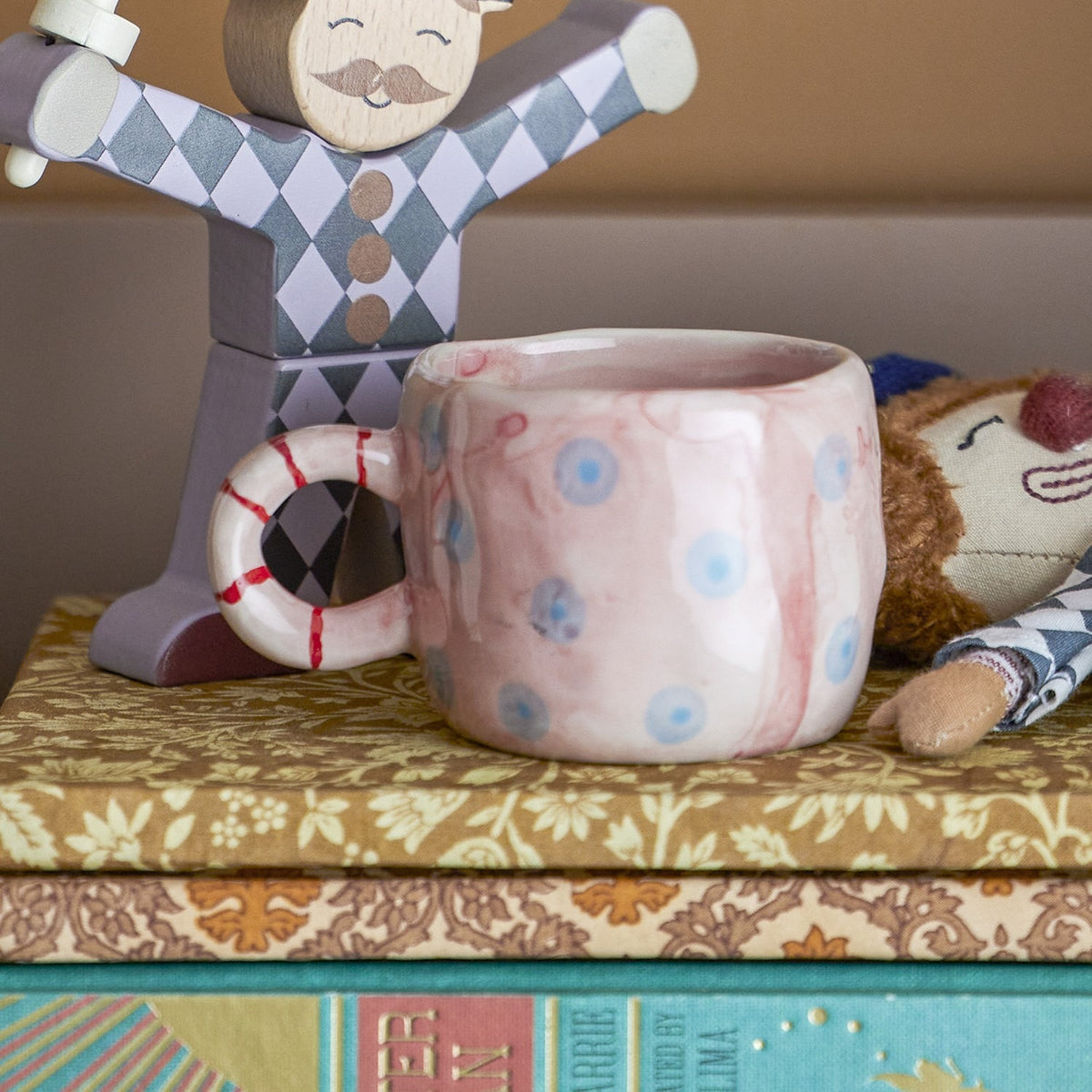 Nini Dotted Stoneware Cup in pink with blue polka dots and a striped ring handle, displayed on books beside wooden and plush toy figures.
