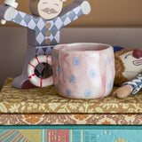 Nini Dotted Stoneware Cup in pink with blue polka dots and a striped ring handle, displayed on books beside wooden and plush toy figures.