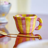 Nini Yellow Striped Stoneware Cup 110ml with a handle, hand-painted in yellow and pink stripes, shown on a reflective surface beside a spoon.