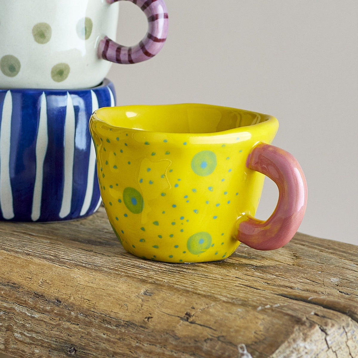 Nini Stoneware Cup 100ml in yellow with hand-painted green and blue dots, a chunky pink loop handle, and an irregular rim, shown on a wooden surface.