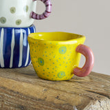 Nini Stoneware Cup 100ml in yellow with hand-painted green and blue dots, a chunky pink loop handle, and an irregular rim, shown on a wooden surface.