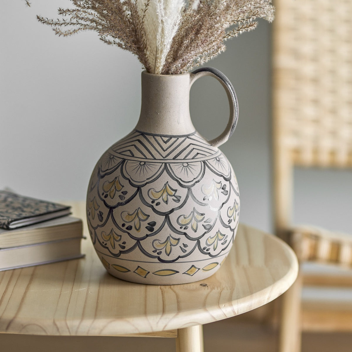 Nadya Round Stoneware Vase 27cm in beige with loop handle, hand-painted chevron, scallop and diamond motifs, shown with dried pampas grass on a wooden table.