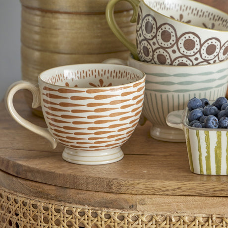 Maple Stoneware Mug 420ml in orange, featuring a footed rounded body with curled loop handle and orange elongated drop patterning, shown among patterned bowls and blueberries on a wooden table.
