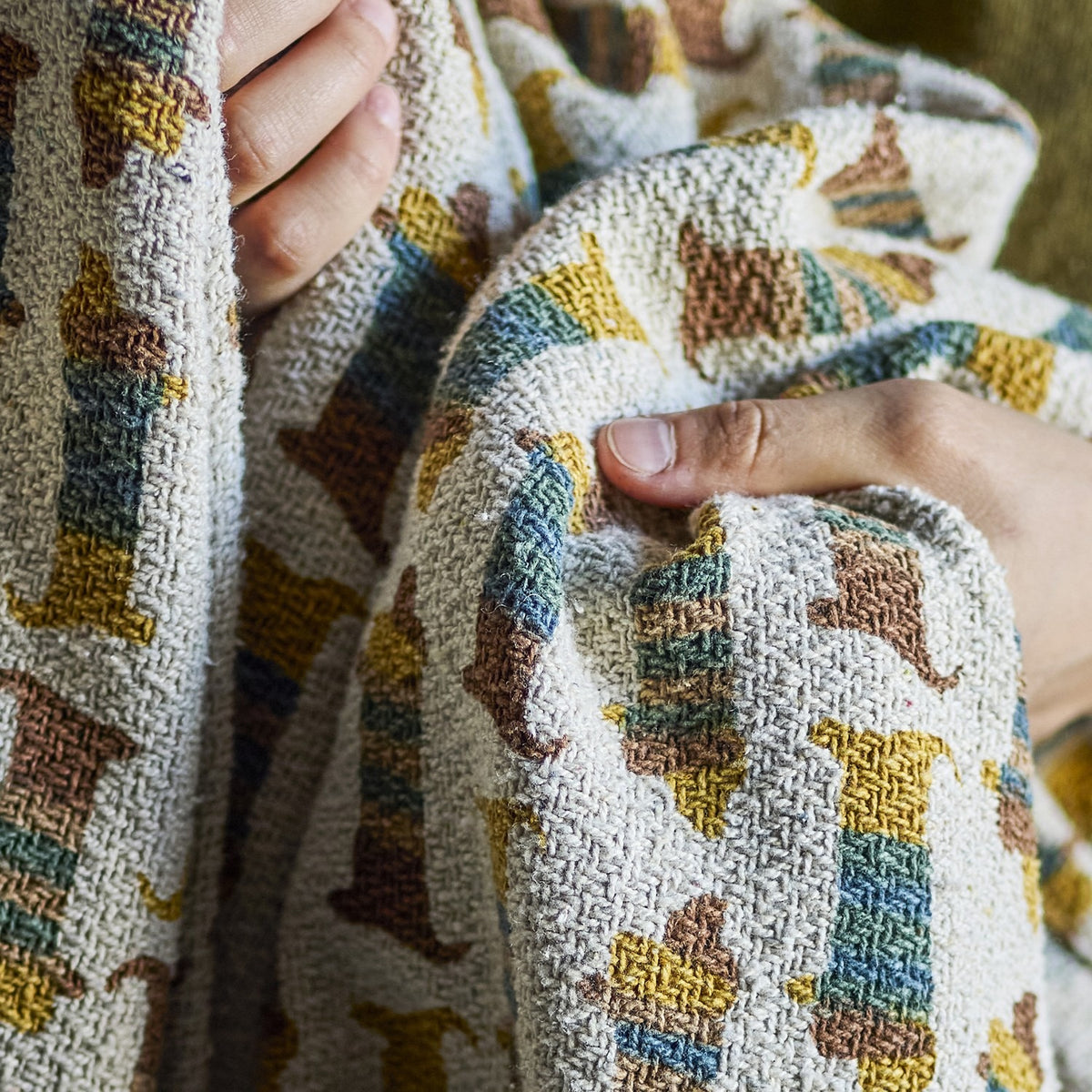 Charlie Cotton Throw in Multi, a woven children's blanket with a repeating sausage dog pattern in mustard, teal and brown, shown held up to display the fabric texture.