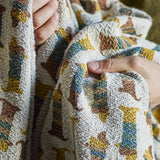 Charlie Cotton Throw in Multi, a woven children's blanket with a repeating sausage dog pattern in mustard, teal and brown, shown held up to display the fabric texture.