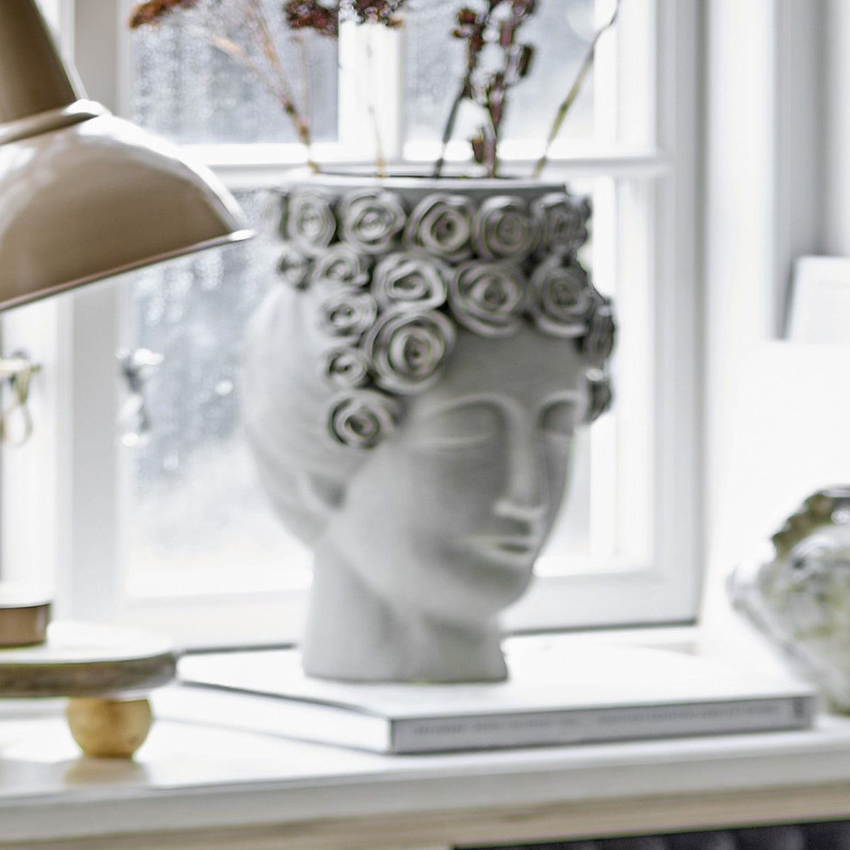 Akira Stoneware Vase 30cm in grey, sculpted as a serene woman's head with a rose wreath rim, holding dried stems on a windowsill beside a lamp and books.
