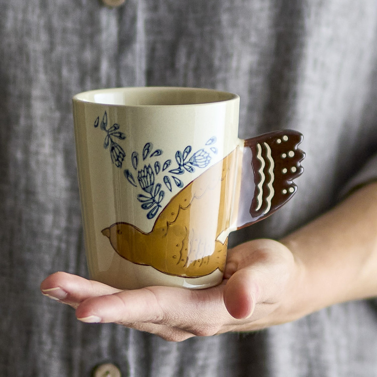 Hezha Stoneware Cup 260ml in cream, hand-painted with a bird in flight forming the brown handle and blue floral sprigs, held in an open palm against a grey backdrop.