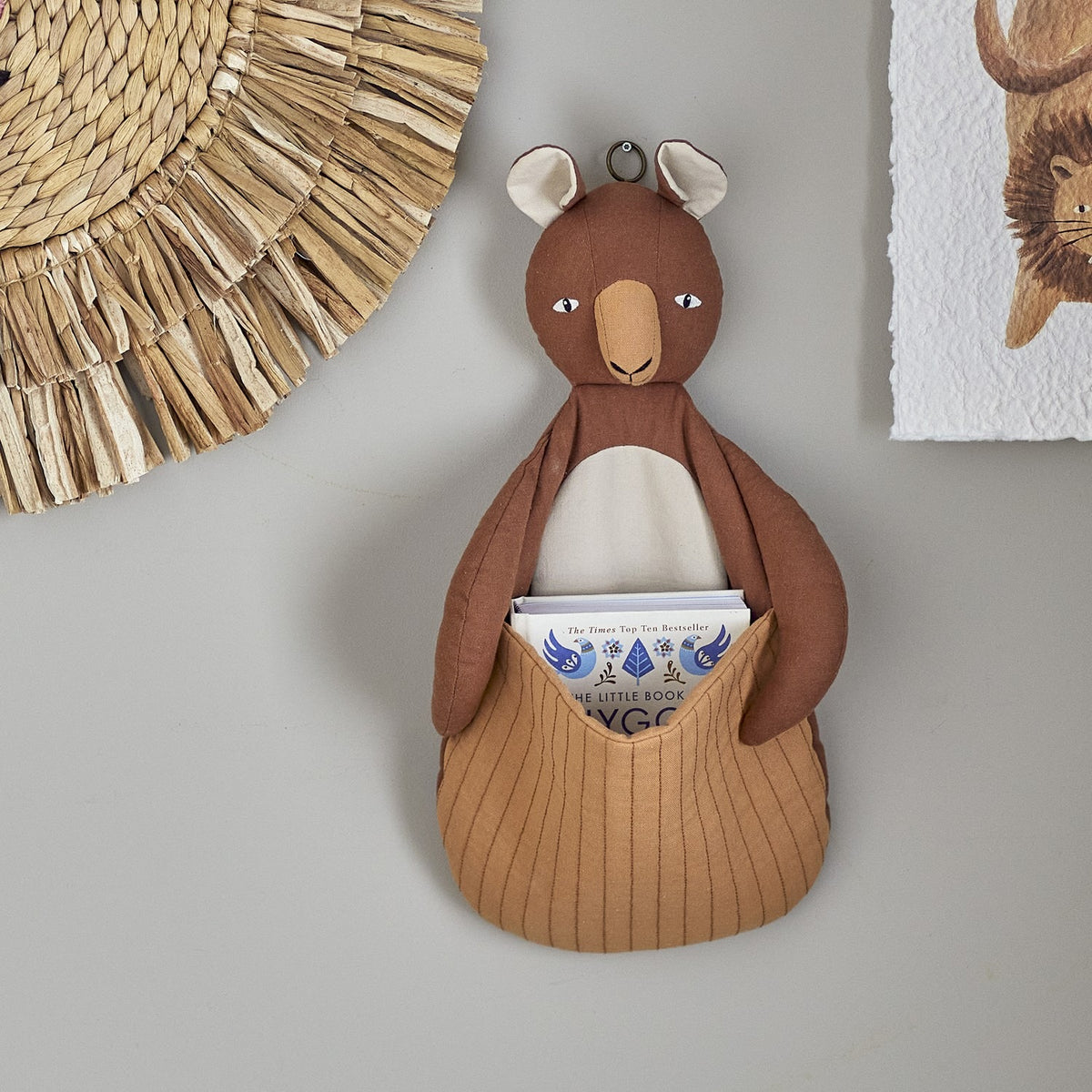 Toto Linen Wall Basket in brown, a kangaroo-shaped fabric storage holder with a quilted pouch holding books, mounted on a grey wall beside woven wall decor.