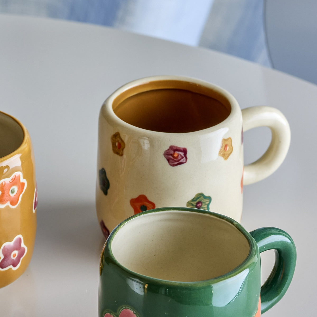 Addy Stoneware Cup 130ml in cream, a small ceramic kids' cup with hand-painted multicoloured flowers and a loop handle, shown beside a green mug on a white table.