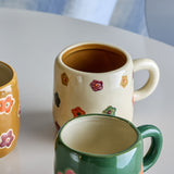Addy Stoneware Cup 130ml in cream, a small ceramic kids' cup with hand-painted multicoloured flowers and a loop handle, shown beside a green mug on a white table.