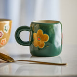 Addy Stoneware Cup 130ml in green with a loop handle and yellow flower design, shown on a table beside a gold spoon and a yellow floral cup.