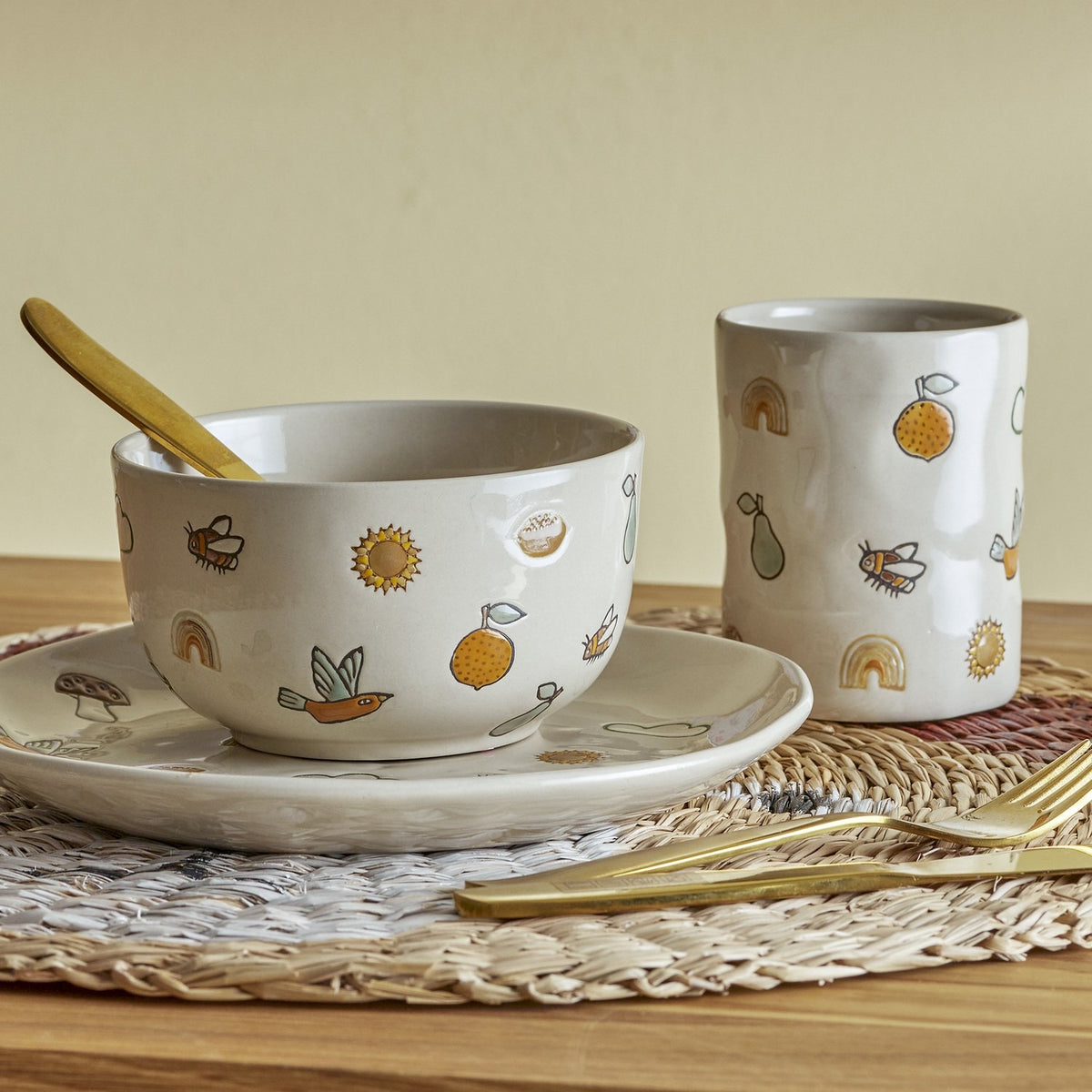 Agnes Stoneware Kids Dinner Set of 3 in cream shown with plate, bowl and cup printed with bees, birds, fruit and rainbow motifs, styled on a woven placemat with gold cutlery.
