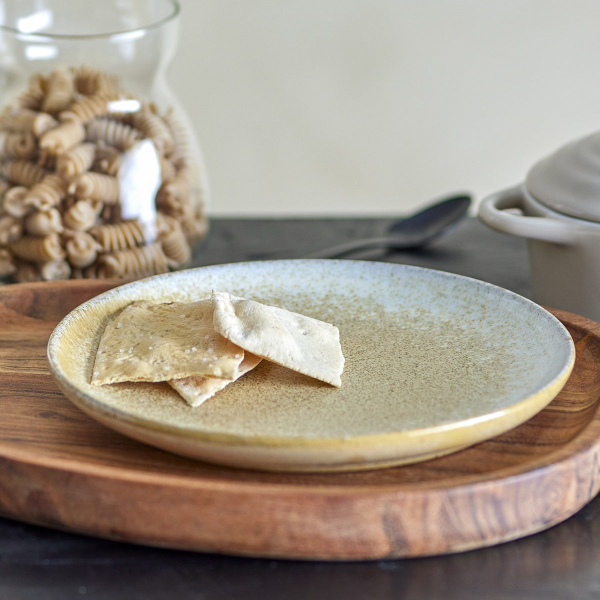 Paula Stoneware beige ceramic breakfast plate with crackers, shown on a wooden board beside a jar of pasta in a kitchen setting.