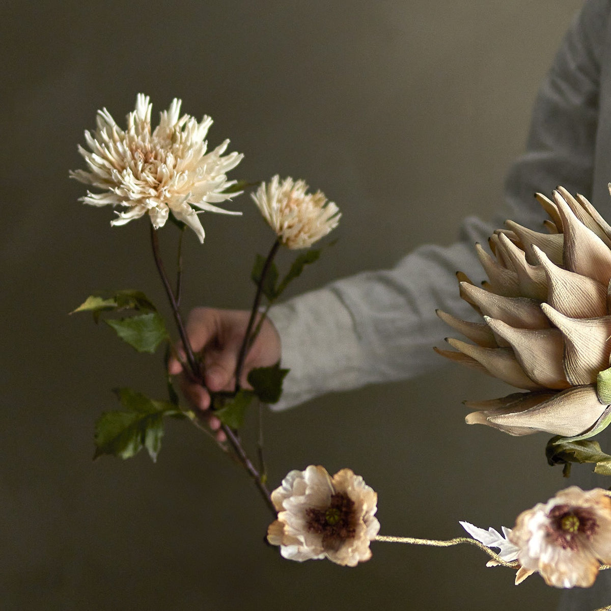 Chrysanthemum Artificial Stem in cream held by hand, showing two layered blooms with pointed petals and serrated leaves on a branching stem.