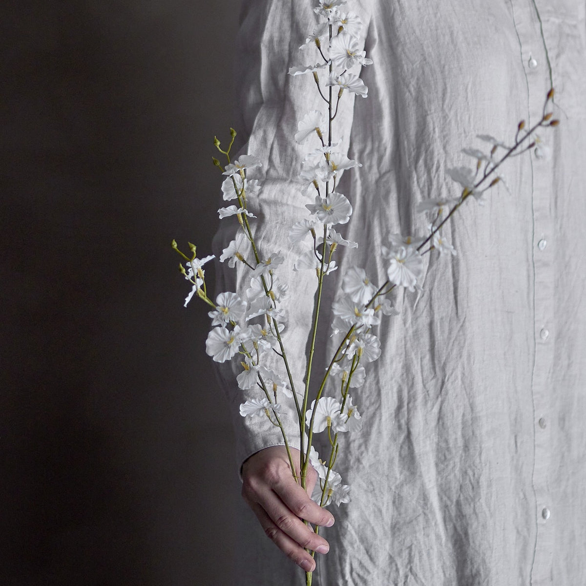 Orchid Artificial Stem 92cm in White, held against a linen shirt, showing slender branches with small white blooms and buds in a lifestyle setting.