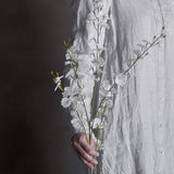Orchid Artificial Stem 92cm in White, held against a linen shirt, showing slender branches with small white blooms and buds in a lifestyle setting.