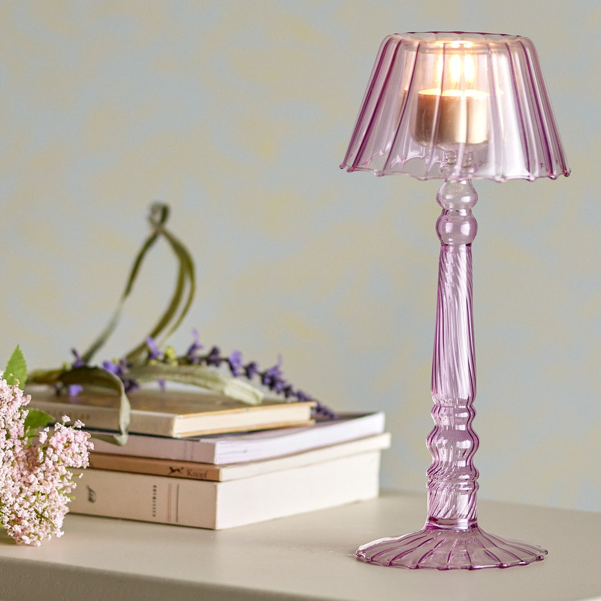 Cisco Glass Tealight Holder 23.5cm in pink, lamp-shaped with fluted scalloped shade and twisted stem, lit with a candle on a shelf beside books and flowers.