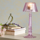 Cisco Glass Tealight Holder 23.5cm in pink, lamp-shaped with fluted scalloped shade and twisted stem, lit with a candle on a shelf beside books and flowers.