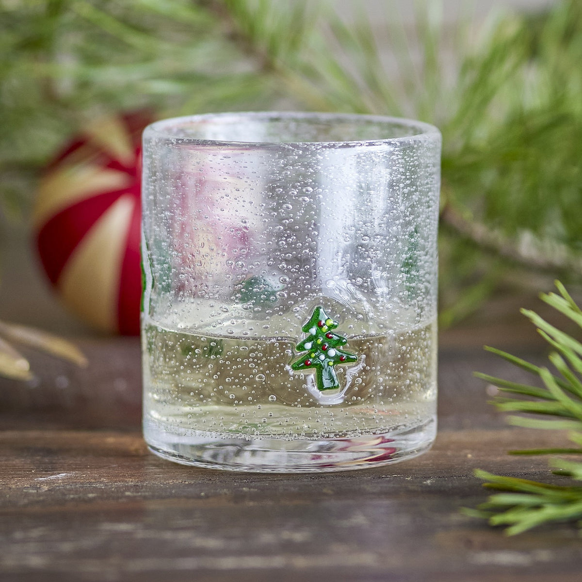 Debra Christmas Tree Tumbler in clear mouthblown glass with fine bubbles and a hand-painted Christmas tree motif, shown filled with sparkling drink among pine branches and a red bauble.