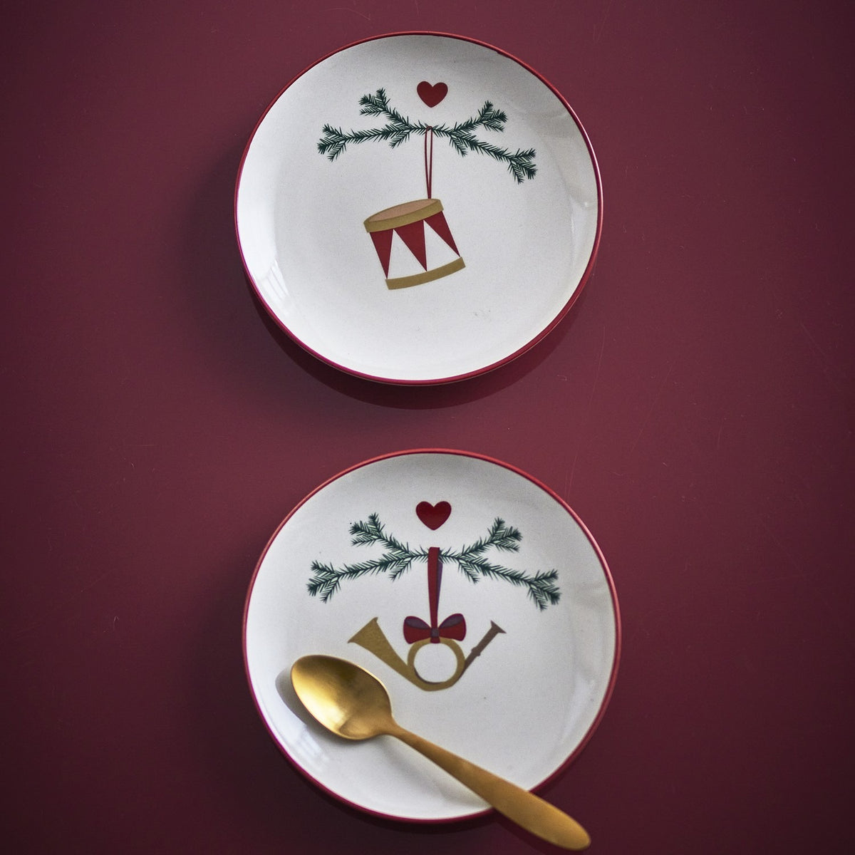Yule Stoneware Christmas Plates Set of 2 in cream, 16cm side plates with red rims, pine sprig and heart motifs, one with a drum and one with a horn, shown on a burgundy background with a gold spoon.