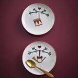 Yule Stoneware Christmas Plates Set of 2 in cream, 16cm side plates with red rims, pine sprig and heart motifs, one with a drum and one with a horn, shown on a burgundy background with a gold spoon.