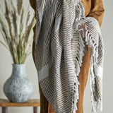 Isnel Brown Striped Cotton Throw draped over a person's arm, showing woven multicoloured stripes, ribbed texture, and a knotted fringe edge.