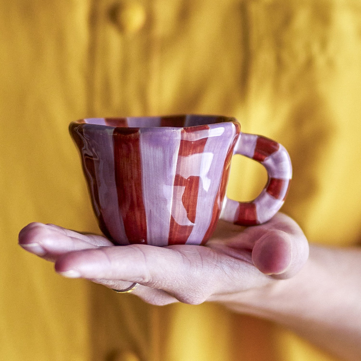 Hand holding the Nini Brown Striped Stoneware Cup, a small 110ml espresso cup with red and lilac vertical stripes and a chunky loop handle, against a mustard yellow backdrop.