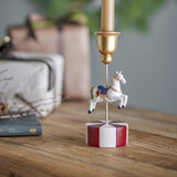 Merry-go-round resin Christmas candle holder with a white carousel horse on a gold rod atop a red and white striped round base, holding a gold candle cup, shown on a wooden table.