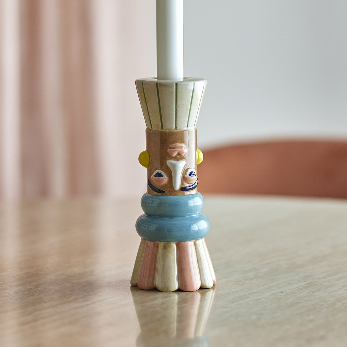 Minio Ceramic Vase 17cm in Multi used as a candle holder, with a face design in cream, tan, blue, pink and yellow, shown holding a white taper candle on a table.