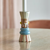 Minio Ceramic Vase 17cm in Multi used as a candle holder, with a face design in cream, tan, blue, pink and yellow, shown holding a white taper candle on a table.