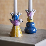 Swift Bird Stoneware Candle Holders, set of 2, with dotted birds beneath leaf-patterned flower cups holding taper candles, one on a yellow cone base and one on a blue double-tier base.