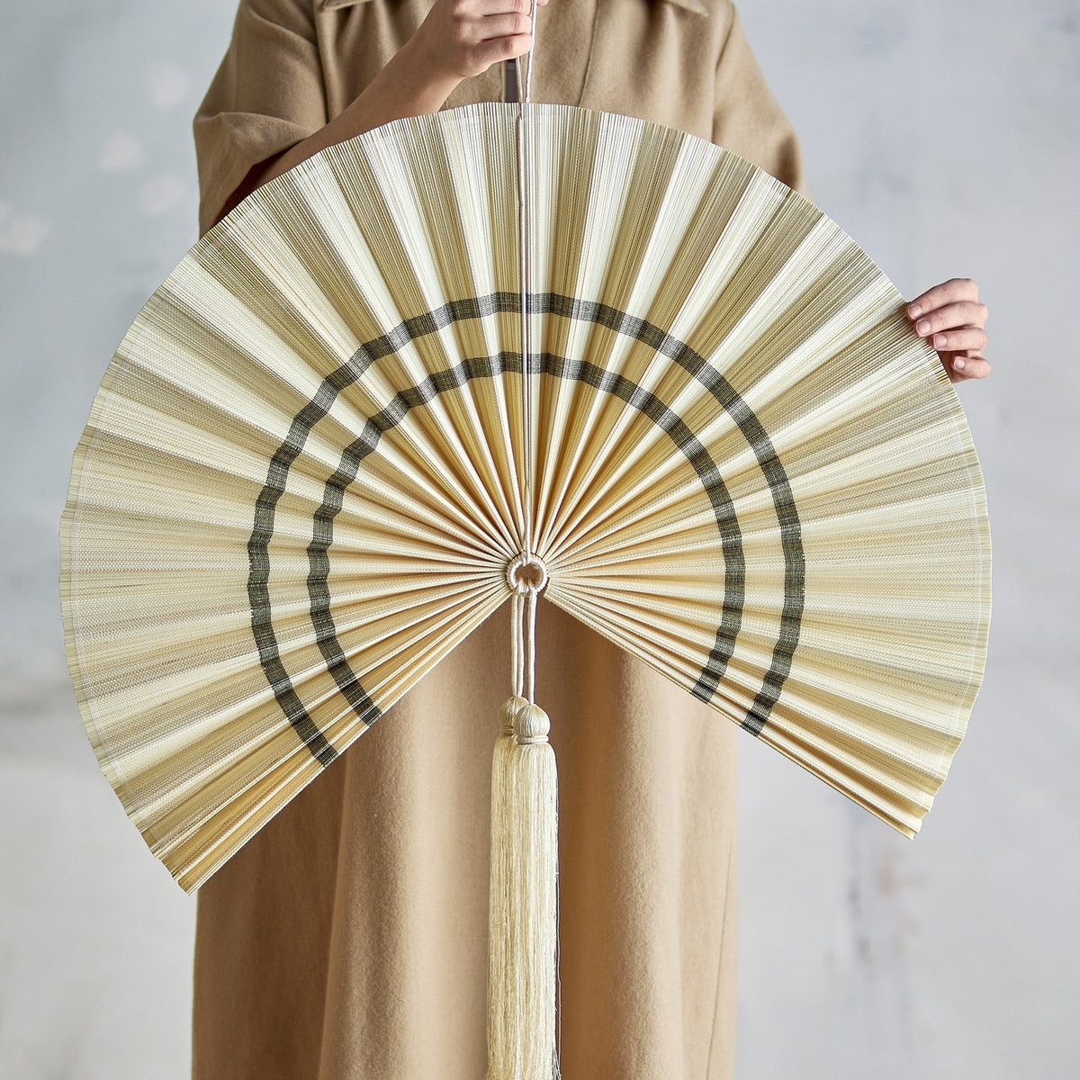 Jaime Bamboo Wall Decor 70cm in natural, a pleated fan-shaped wall art piece with contrasting curved bands and a long tassel, held open to show scale against a plain background.