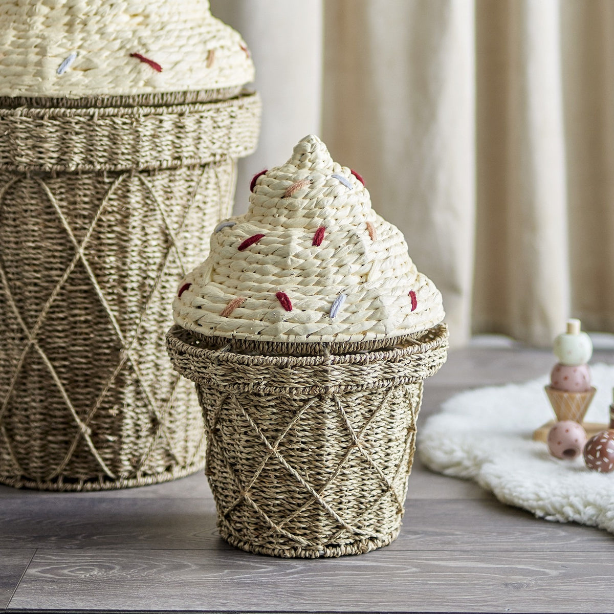 Cillie 41cm Natural seagrass basket with lid, ice-cream cone shape, criss-cross woven base, domed swirl lid with yarn sprinkles, shown beside a larger matching basket in a nursery.