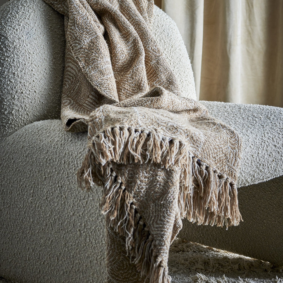 Okapi Cotton Throw in beige with woven wavy-line pattern and fringed edges, draped over a bouclé armchair in a living room setting.