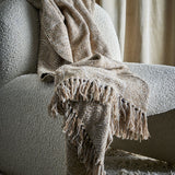 Okapi Cotton Throw in beige with woven wavy-line pattern and fringed edges, draped over a bouclé armchair in a living room setting.