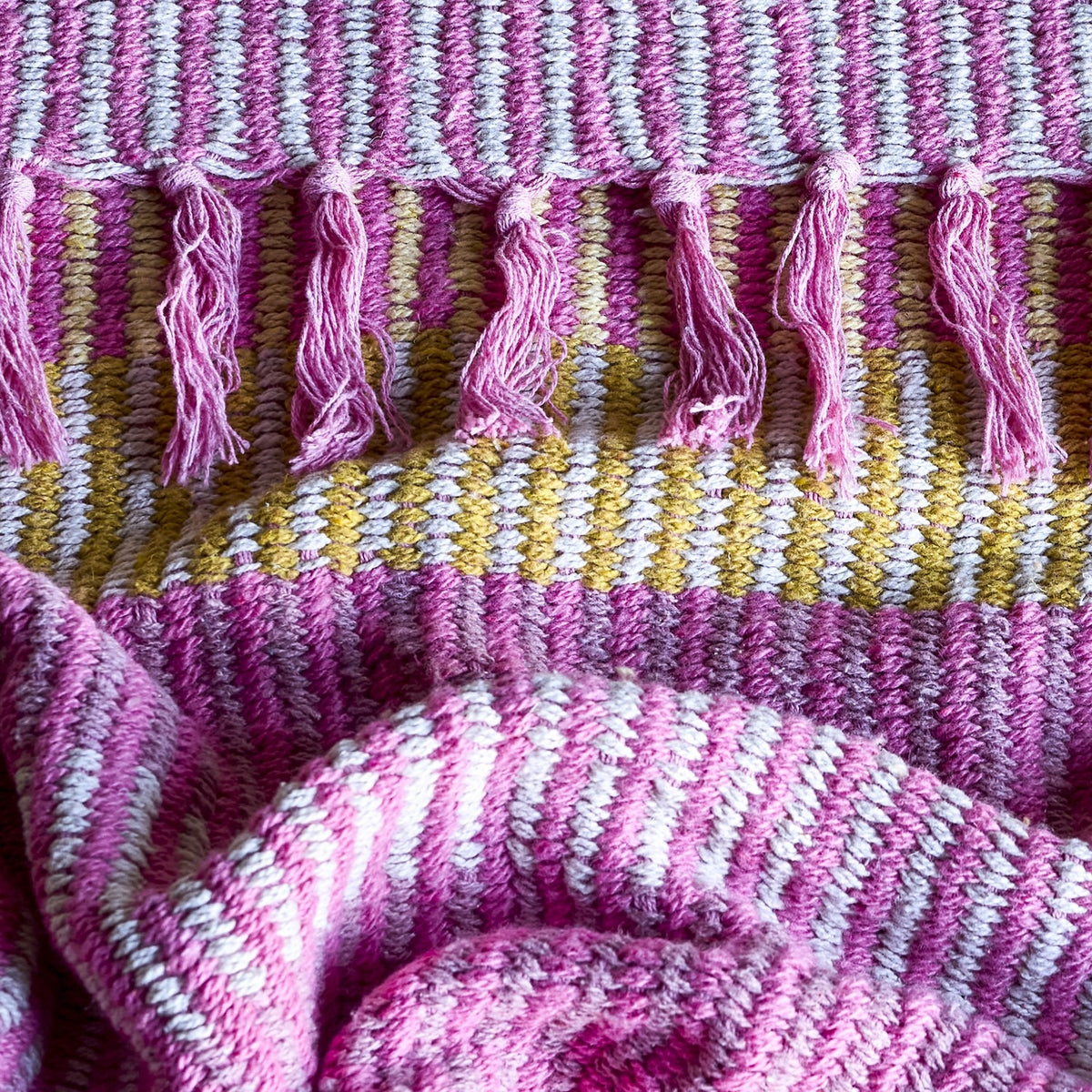 Isnel Striped Throw in pink, close-up of the textured woven fabric with striped bands in pink, mustard and white, finished with knotted fringe tassels.