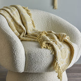 Pratello Throw in yellow with a woven floral leaf pattern and fringed edges, draped over a cream bouclé armchair.