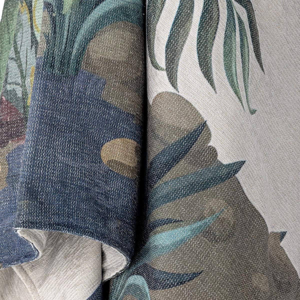Peacock Throw in green, close-up of folded woven fabric showing palm fronds, leaves and stone shapes in muted green, grey and blue tones.