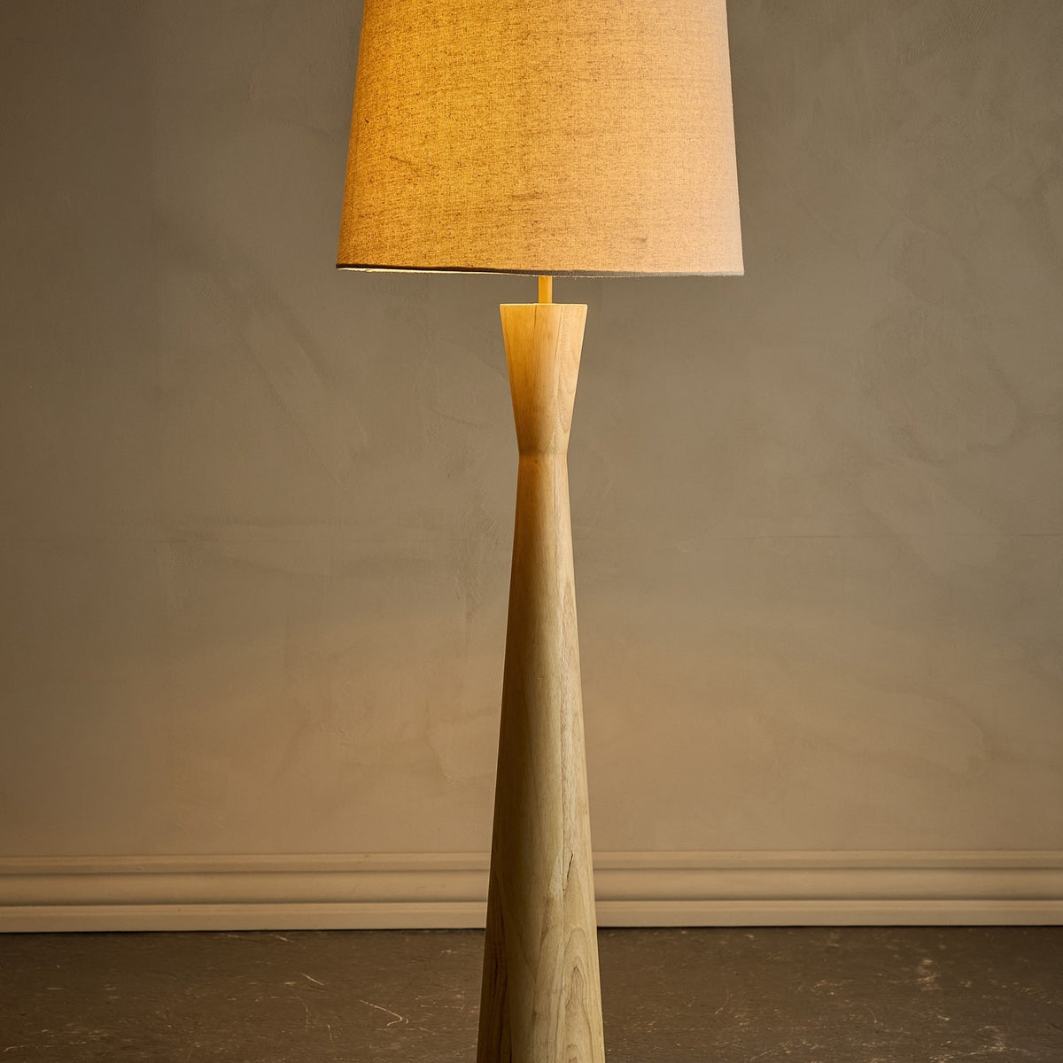 Leonor Wooden Floor Lamp with Linen Shade in Natural, showing the tapered rubberwood column with pinched waist and tan linen drum shade, set against a plain wall.